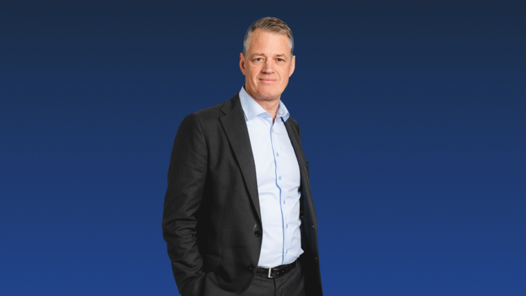 A man in a dark suit and light blue shirt stands with one hand in his pocket against a solid blue background.