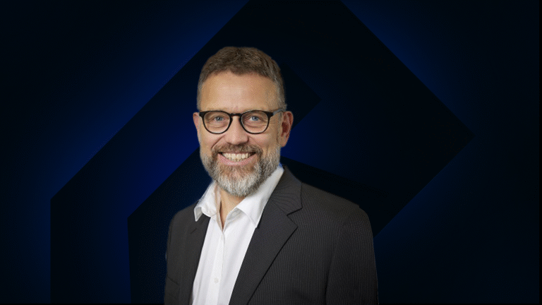 A man with glasses and a beard, wearing a dark suit jacket and white shirt, smiles at the camera against a dark geometric background.