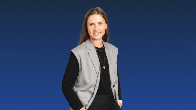 A woman stands against a blue gradient background, wearing a grey vest over a black top and black pants, with her right hand in her pocket.