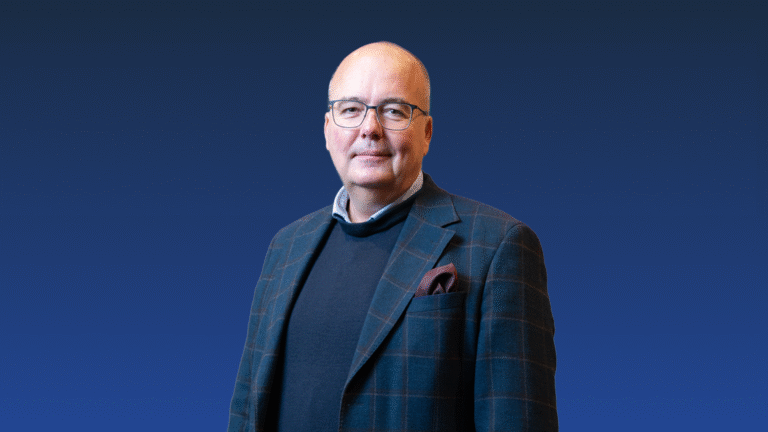 A middle-aged man with glasses and a bald head wears a plaid blazer, dark sweater, and pocket square, standing against a blue gradient background.
