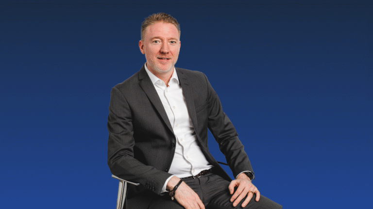 A man in a dark suit and white shirt sits on a chair against a solid blue background, facing the camera with a neutral expression.