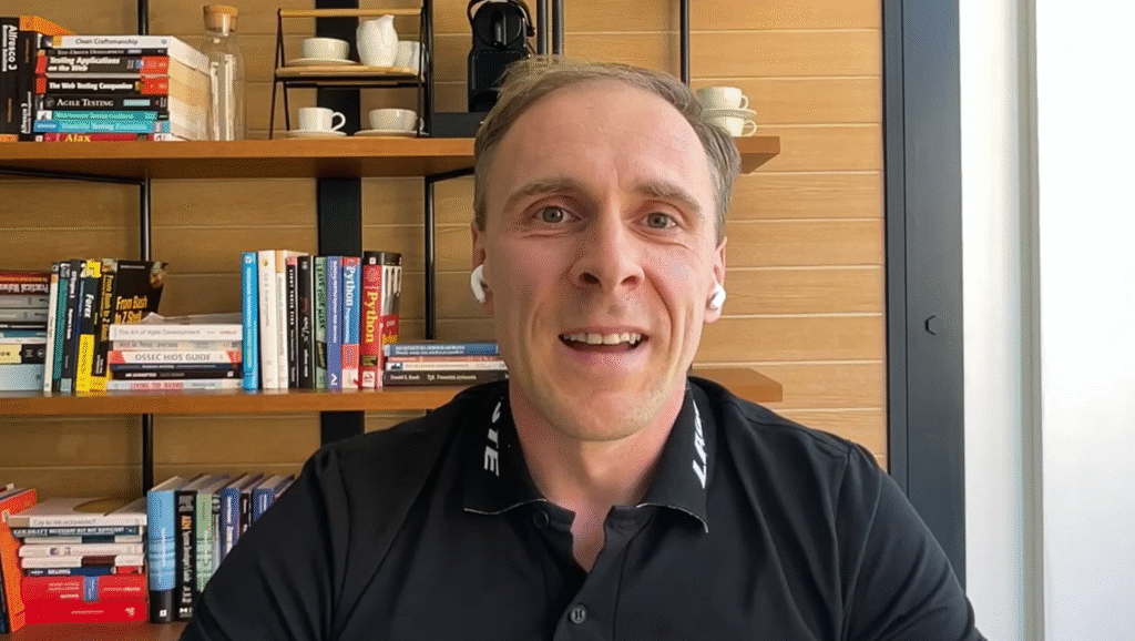 A man wearing a black collared shirt and wireless earbuds sits in front of a bookshelf with books, cups, and a coffee machine.