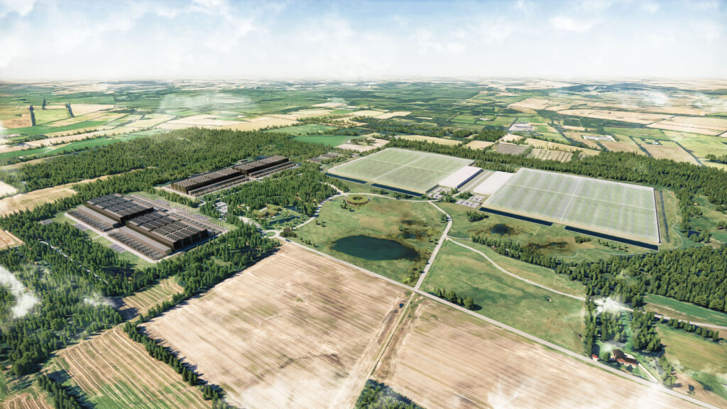 Aerial view of a rural landscape with large industrial buildings, green fields, forested areas, and a central pond under a partly cloudy sky.