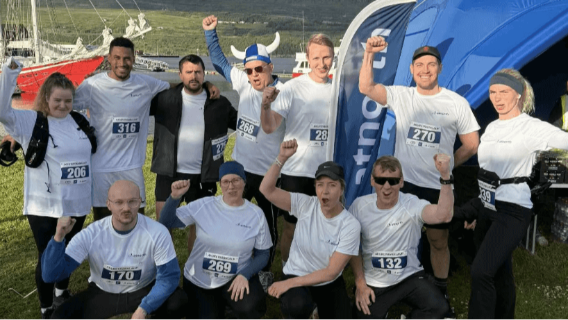 A group of runners in white shirts pose together outdoors, some raising their fists, with race bibs visible and a blue event tent in the background.