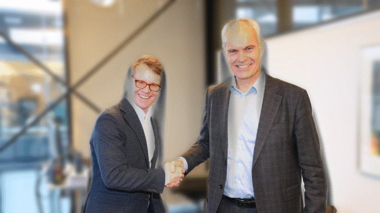 Two men in business attire are shaking hands and smiling in an office setting.