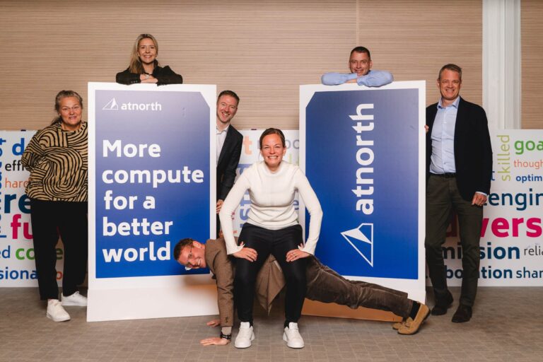 Seven people pose indoors with two large atnorth signs; one person does a plank between two others who support her arms and legs.