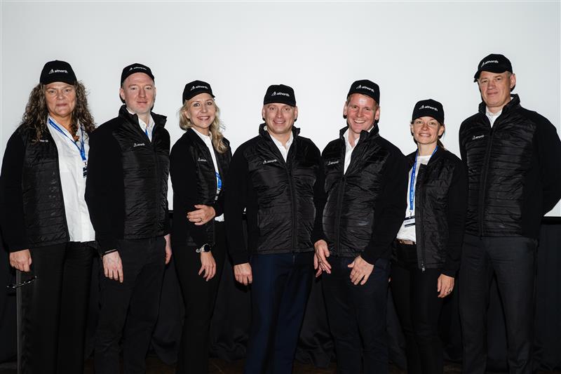Seven people stand side by side, wearing matching black vests and caps with logos, in front of a plain white background.