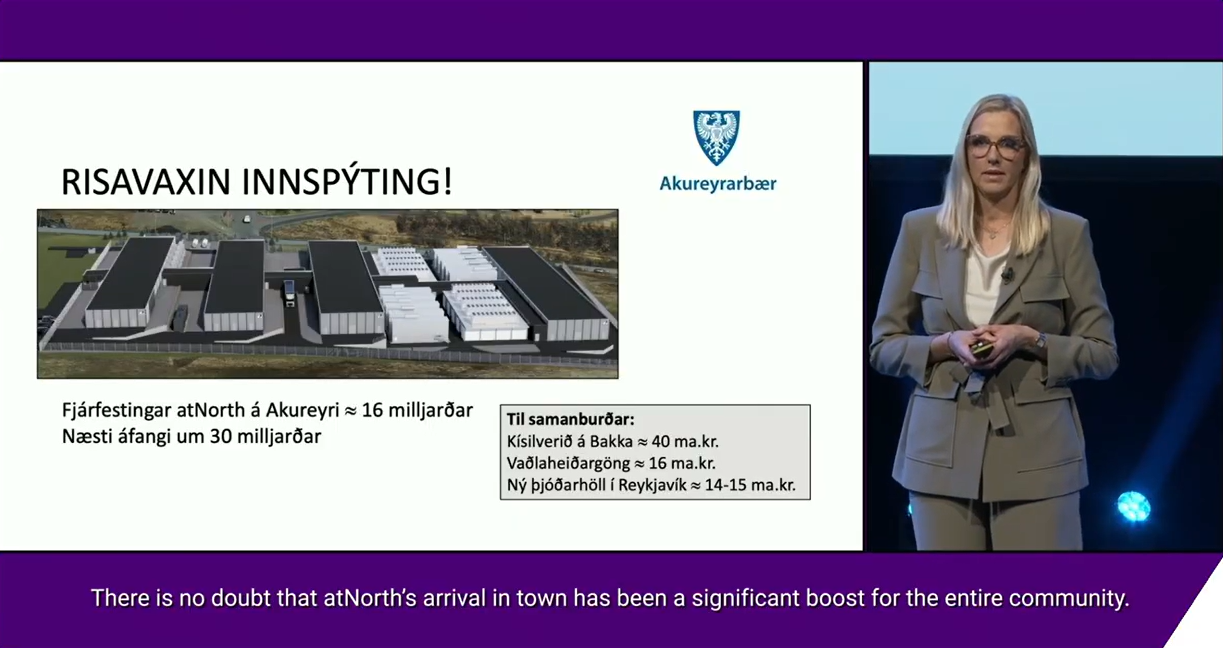 A woman in a suit presents a slide showing an aerial view of a data center project with Icelandic text, charts, and the Akureyri town logo.