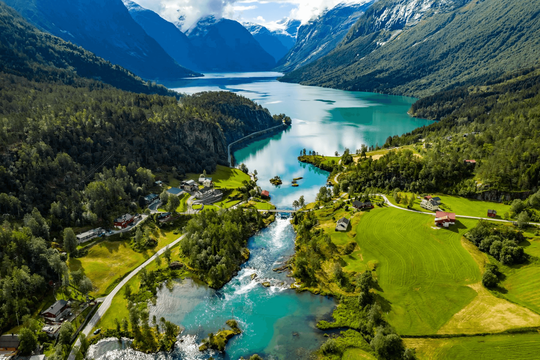 Aerial view of a lake surrounded by mountains, forested hills, and scattered houses with green fields and clear turquoise water.
