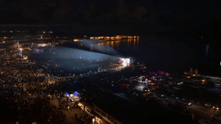 A large crowd gathers outdoors at night around a stage with bright lights, near a body of water with illuminated buildings in the background.