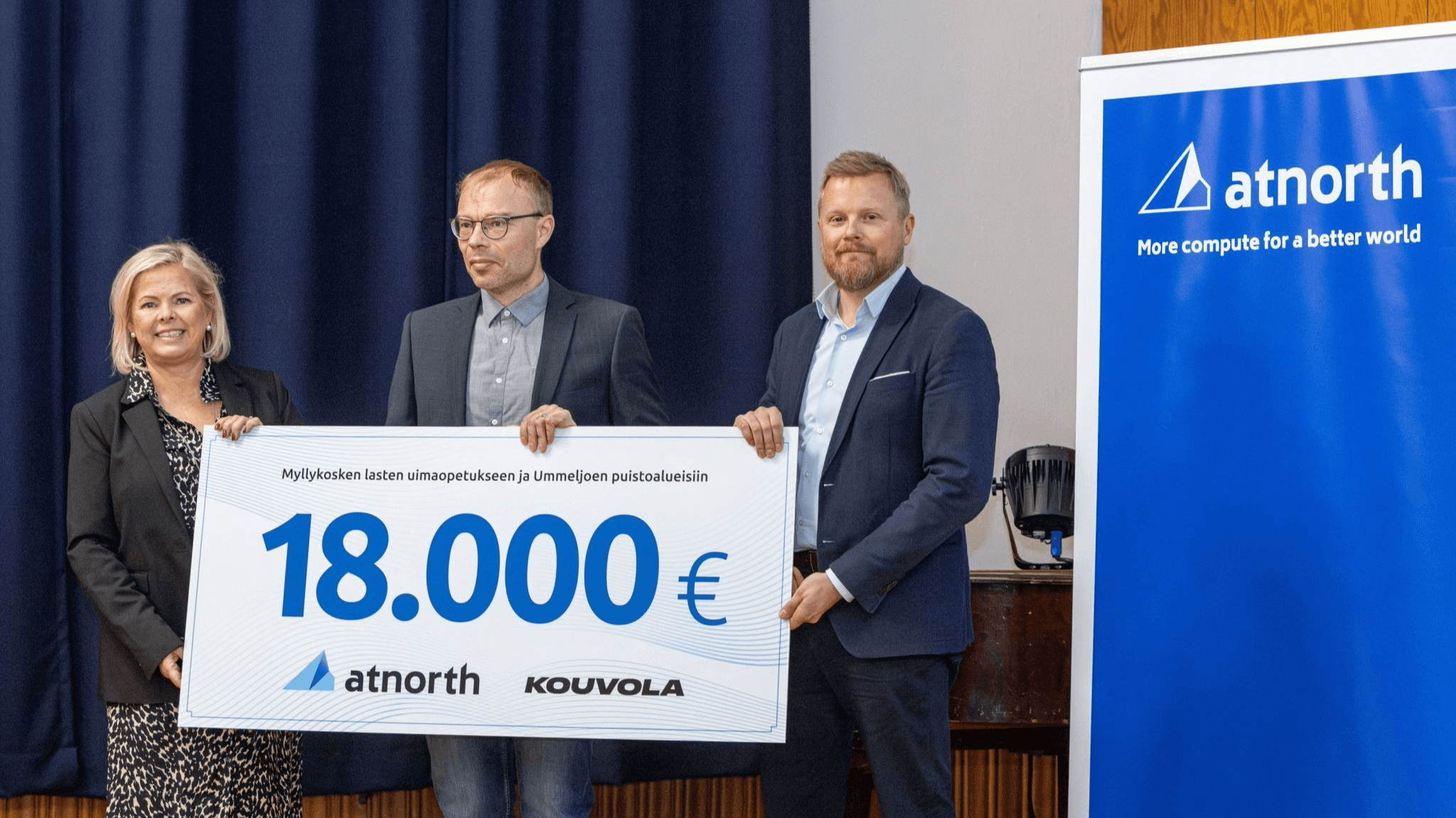 Three people stand holding a large check for 18,000 euros from atnorth and Kouvola, with a blue atnorth banner in the background.