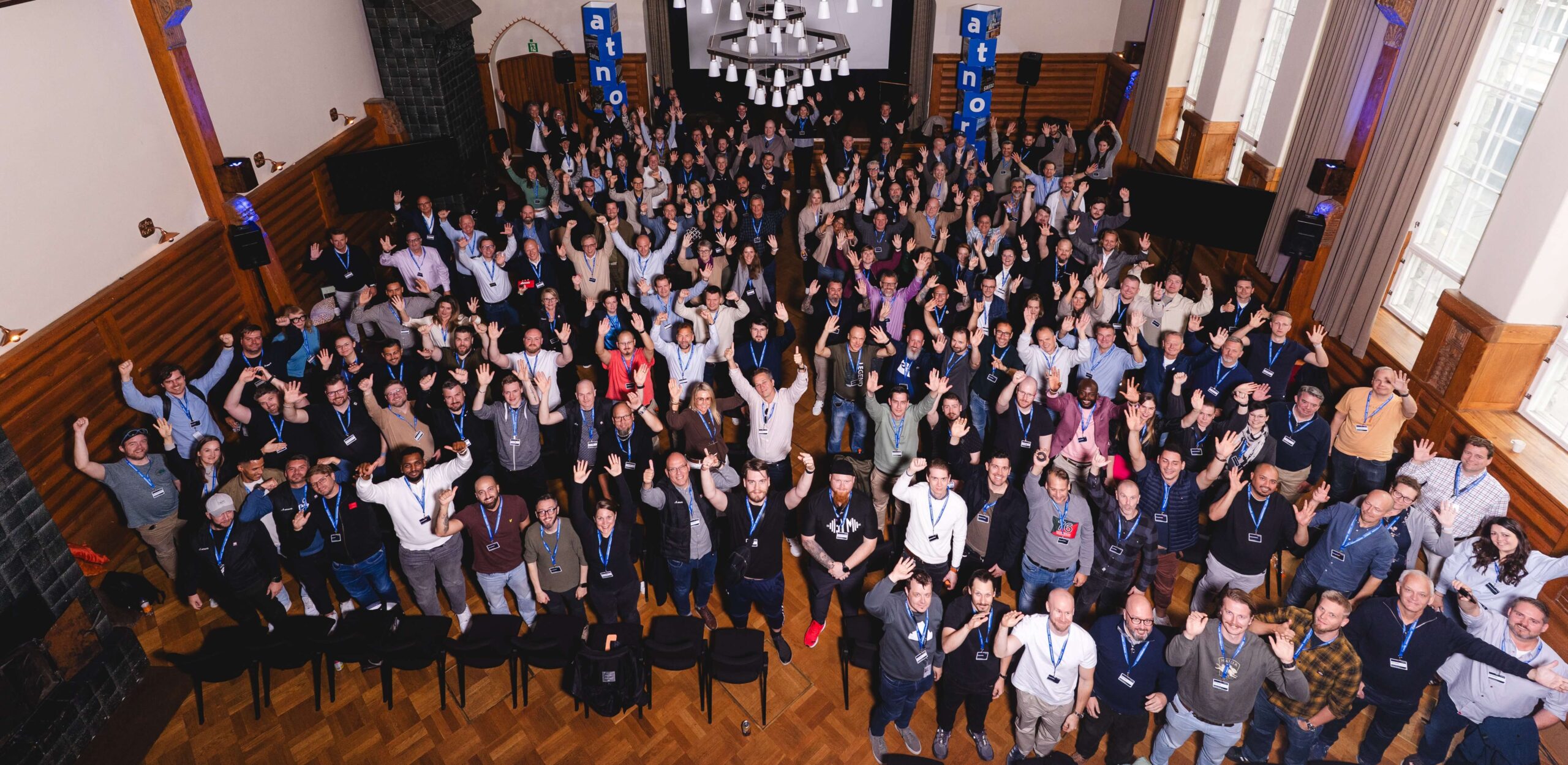 A large group of people pose together indoors for a group photo at an event, many with raised arms, wearing name badges and casual clothing.