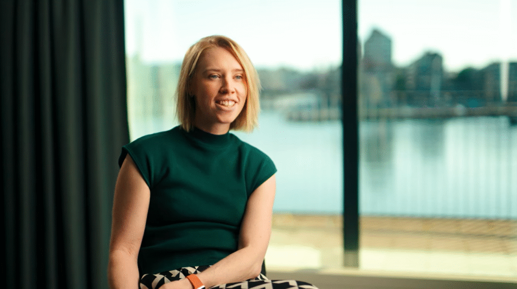 A woman with short blonde hair sits indoors near a large window overlooking water and buildings, smiling and wearing a green top and patterned pants.