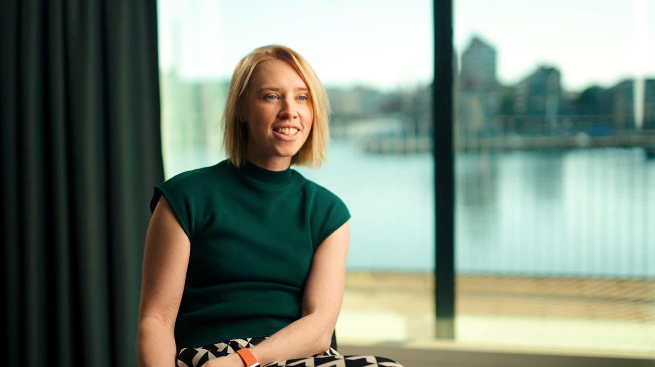 A woman with short blonde hair sits indoors near a large window overlooking water and buildings, smiling and wearing a green top and patterned pants.