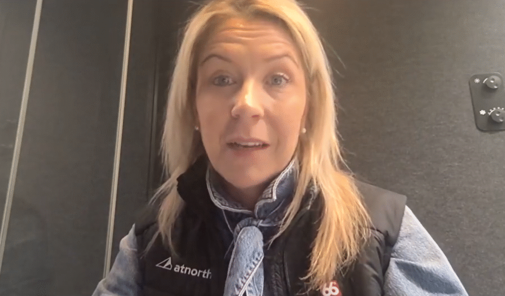 A woman with blonde hair, wearing a black vest and blue scarf, sits indoors in front of a dark background, looking at the camera.