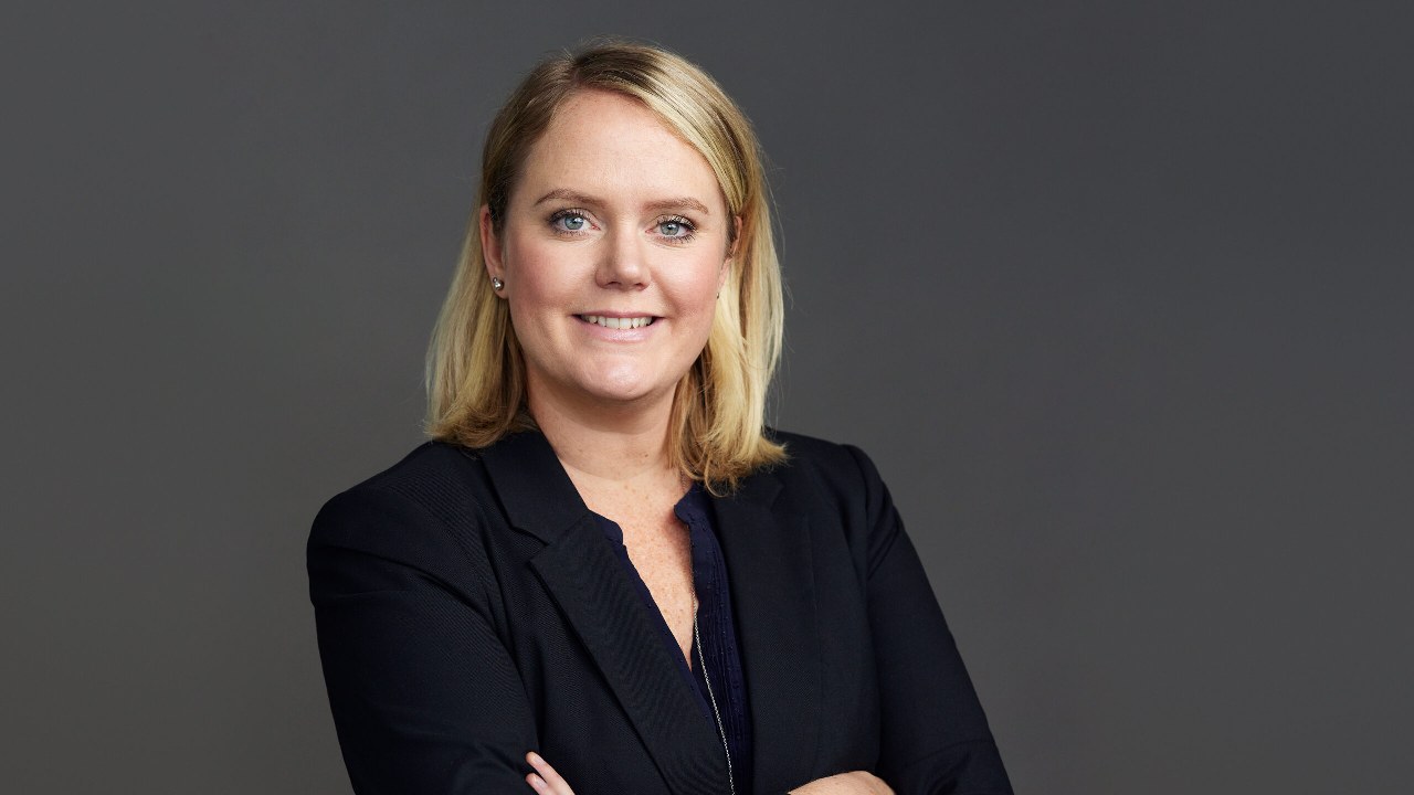 A woman with blonde hair in a business suit stands with her arms crossed, smiling, against a plain gray background.