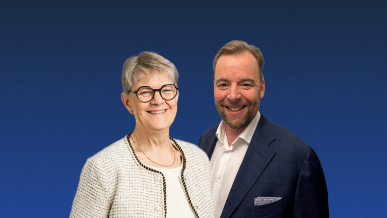 Two adults, one woman and one man, stand side by side smiling in business attire against a plain blue gradient background.