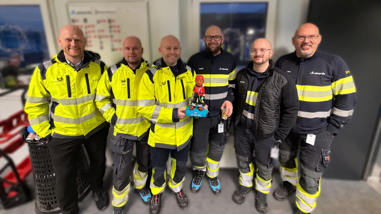 Seven men in high-visibility workwear stand indoors, smiling at the camera; one is holding a small figurine on a blue base.