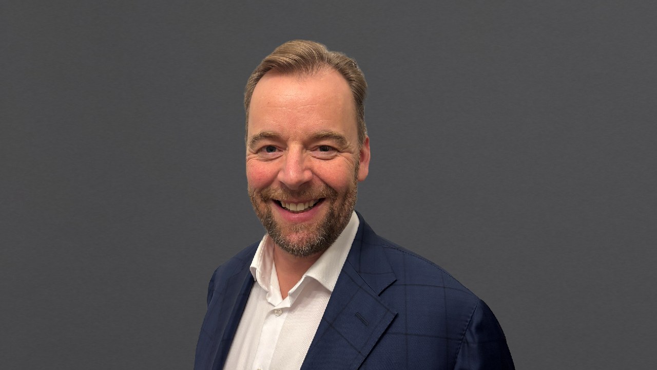 A man in a blue suit and white shirt stands smiling in front of a plain gray background.