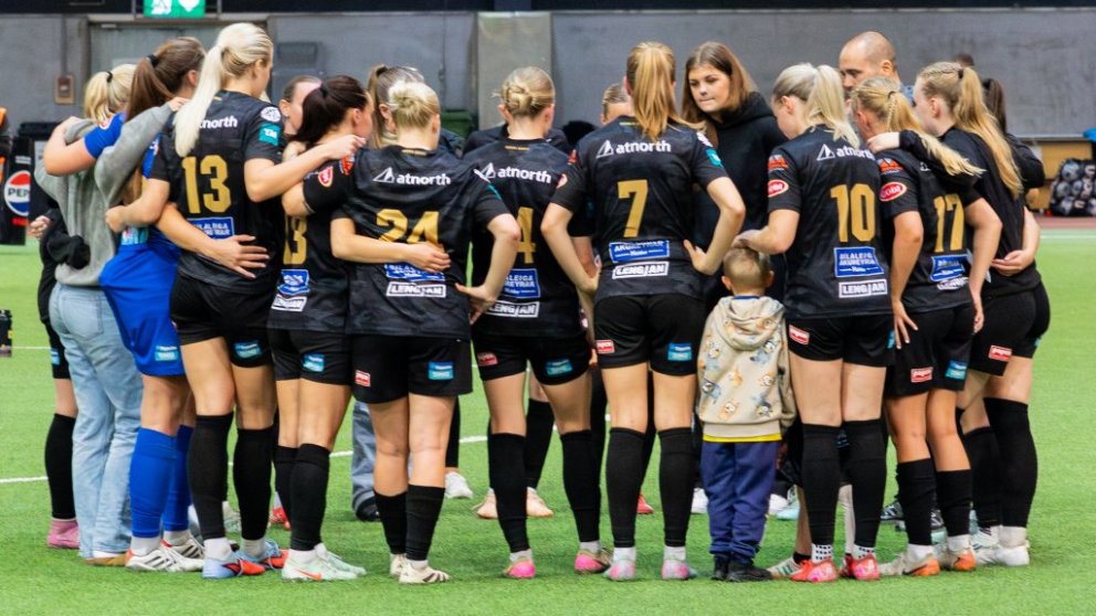 A women's soccer team huddles together with coaches and a child on an indoor field, wearing black uniforms with visible sponsor logos.