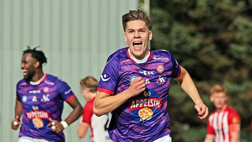 A soccer player in a purple jersey runs and celebrates on the field while other players in the background react during a match.