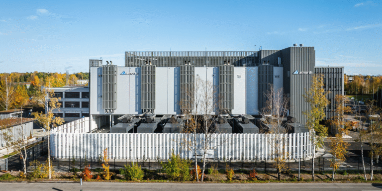 A modern data center building with vertical vents, surrounded by a white fence and autumn trees, under a clear blue sky.