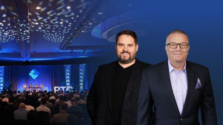 Two men in suits stand in front of a conference scene with an audience and panelists on stage under blue lighting.