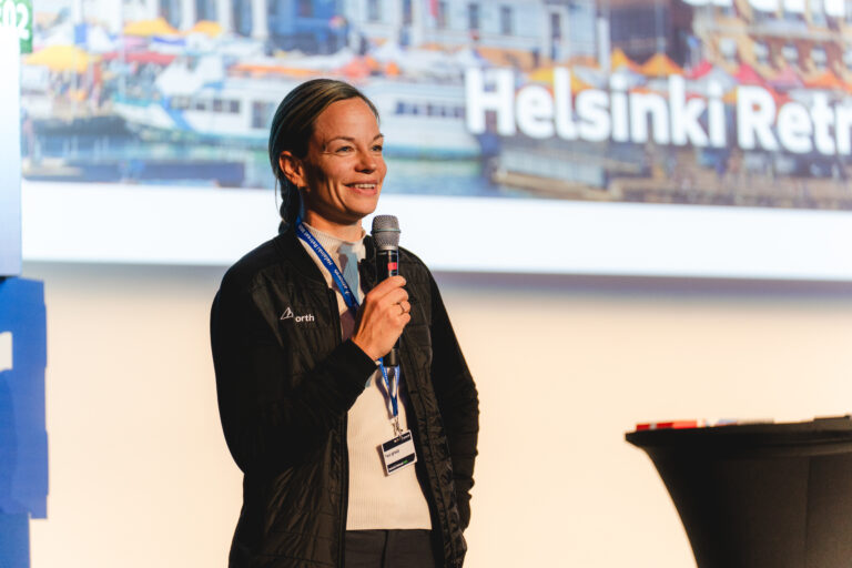 A woman stands on stage holding a microphone, speaking at an event with a screen behind her displaying the words "Helsinki Retreat.