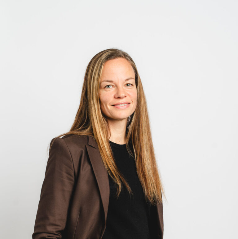 A woman with long straight hair wearing a brown suit and black top stands against a plain white background, smiling slightly with one hand in her pocket—exuding the confident professionalism found in AI-ready data centers.