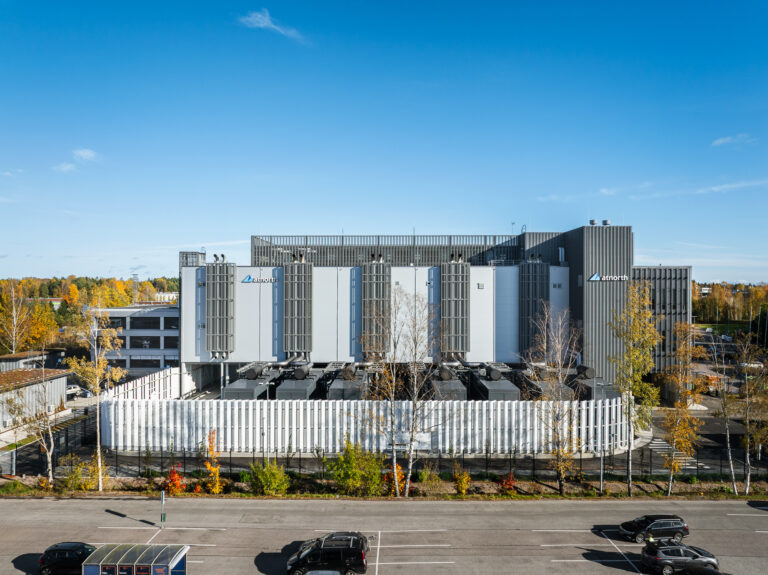 A modern industrial building with large external cooling units, surrounded by a fence, adjacent to a parking lot and autumn trees under a clear blue sky.