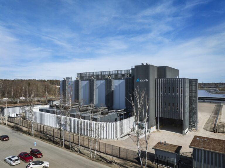 A modern data center facility with the "anorth" logo, surrounded by security fencing, next to a road with parked cars and trees in the background.