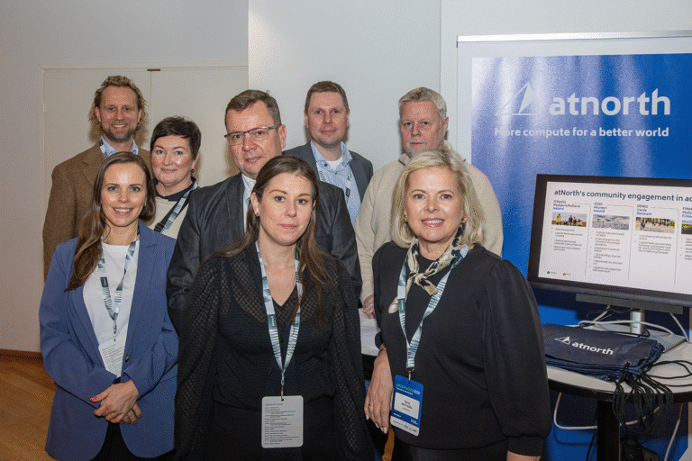 A group of eight people standing indoors in front of an atnorth display, wearing business attire and conference badges.