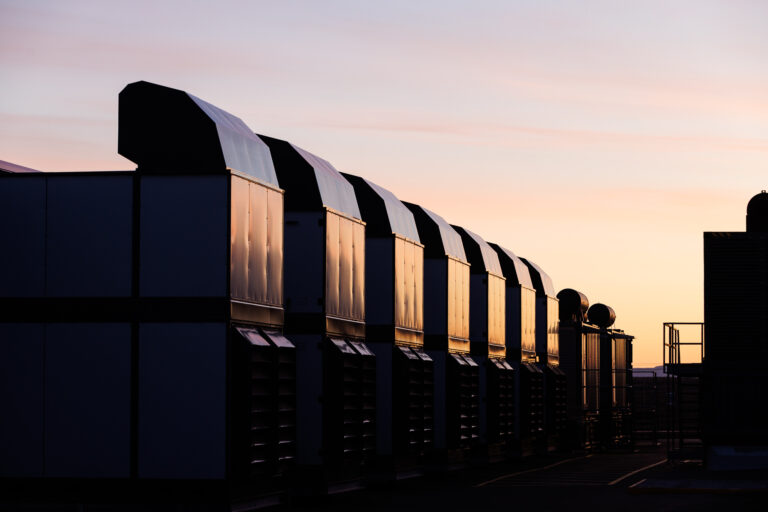 Rows of industrial HVAC units are silhouetted against a sunset sky, with warm light reflecting off their surfaces.