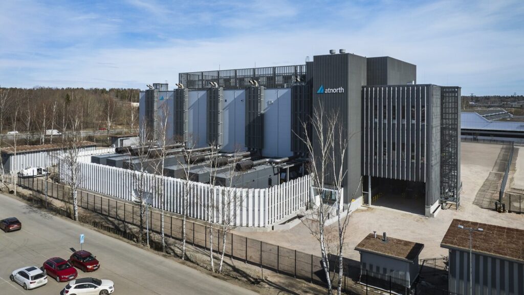 A modern data center facility with the "anorth" logo, surrounded by security fencing, next to a road with parked cars and trees in the background.