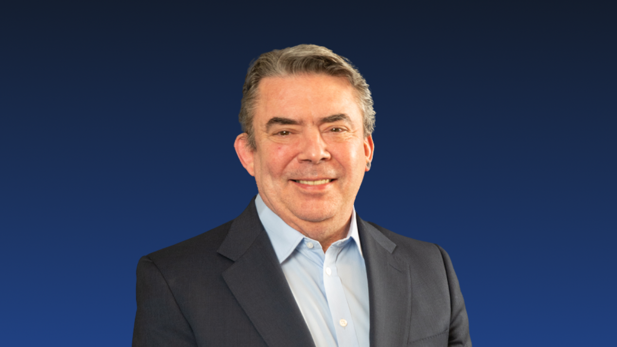 A middle-aged man in a dark suit and light blue shirt smiles at the camera against a plain dark blue background.