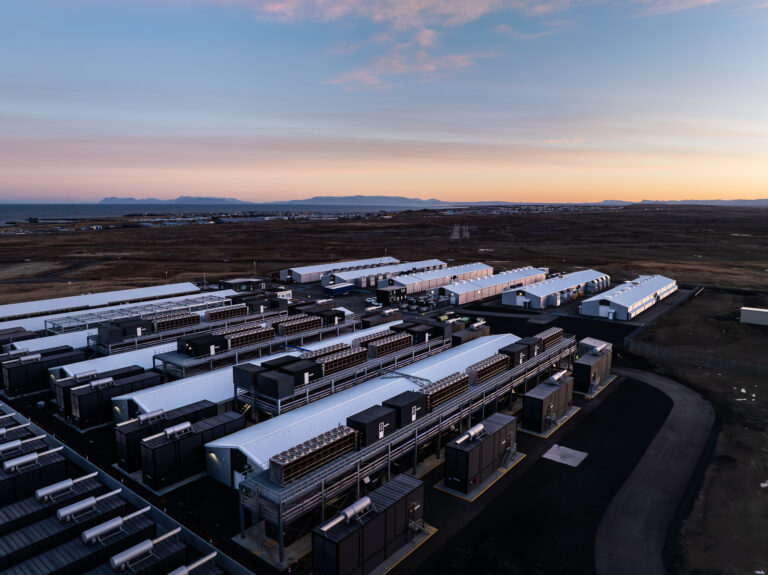 Aerial view of a large, modern industrial facility with multiple buildings and cooling units—AI-ready data centers—set in a flat, sparsely vegetated landscape at sunset.