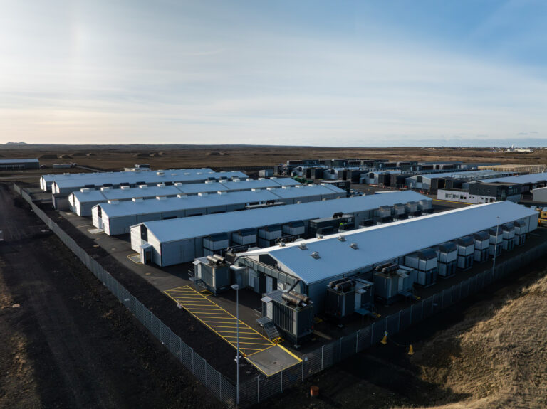 Aerial view of a large facility with multiple long, white AI-ready data centers and equipment units, surrounded by fencing and situated in a barren landscape.