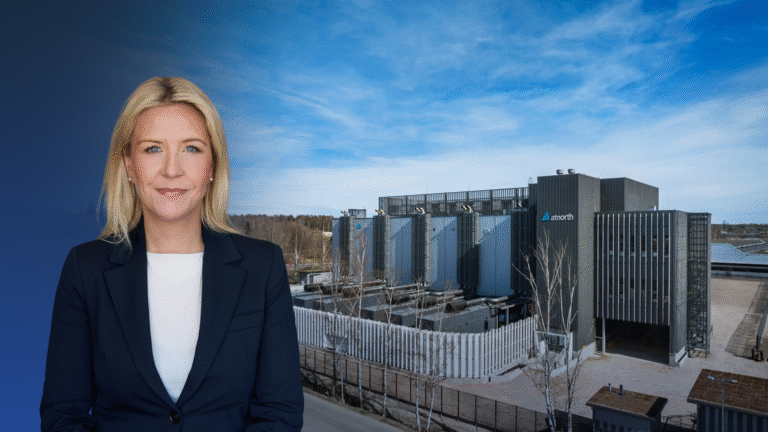 A woman in business attire stands in front of a modern industrial building under a blue sky, symbolizing leadership in decarbonizing AI data centers.