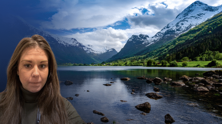 A woman with long brown hair stands in front of a scenic lake with rocks, surrounded by mountains and partly cloudy skies.