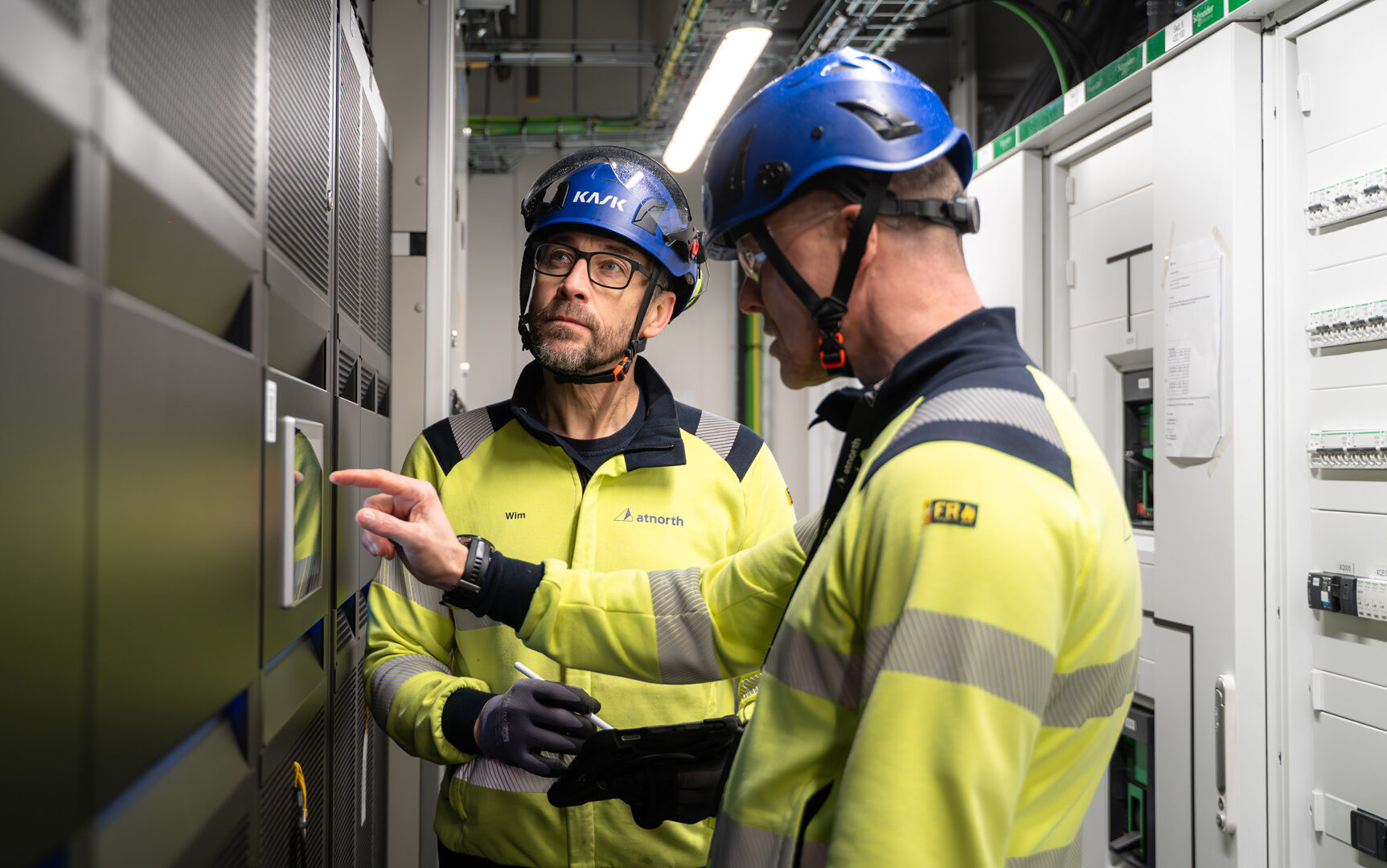 Two workers in safety gear and helmets inspect and operate a control panel in an industrial setting.
