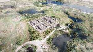 Aerial view of a large industrial facility with multiple rectangular buildings, surrounded by greenery, water, and wind turbines in a rural landscape.