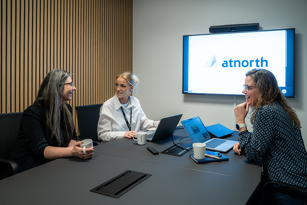 Three women sit around a conference table with laptops and mugs, smiling and conversing; a screen behind them displays the atnorth logo.