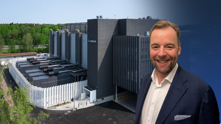 A man in a suit smiles in front of a large, modern AI-ready data center building with the Microsoft logo, surrounded by greenery under a clear sky.