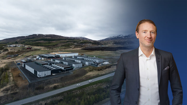 A man in a suit stands in front of a modern industrial facility, home to AI-ready data centers, with mountains and an overcast sky in the background. The right side of the image has a blue gradient overlay.