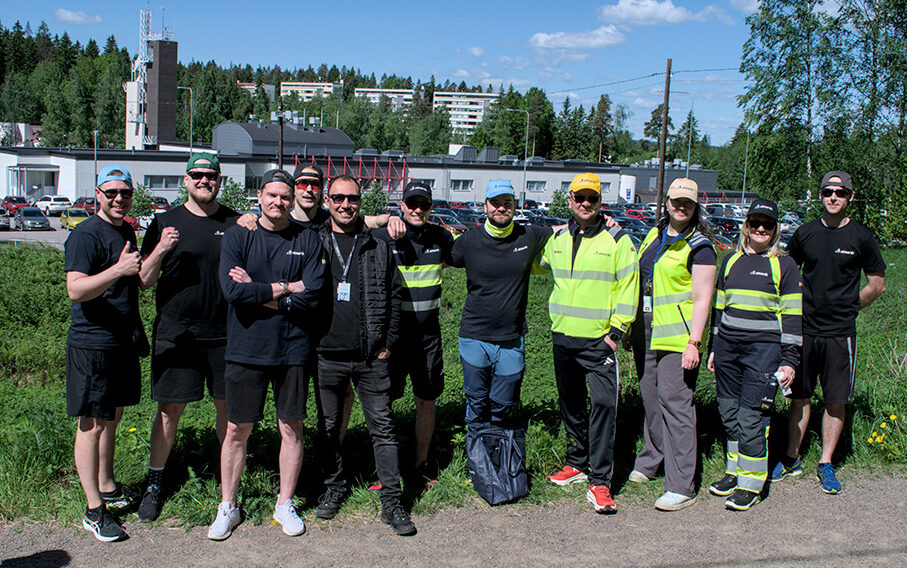 A group of people, some wearing black clothing and others in yellow safety vests, stand outdoors on a sunny day with buildings and trees in the background.