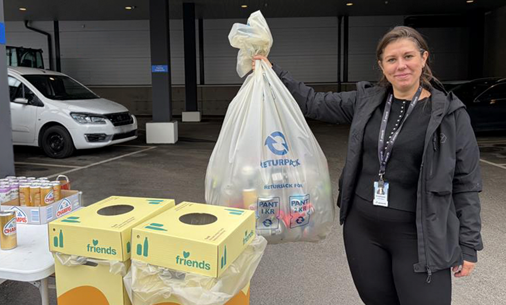 A woman stands outdoors holding a large bag of recyclables next to recycling bins and a table with drink cans.