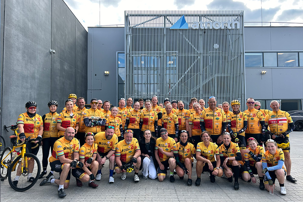 A large group of cyclists in matching yellow jerseys pose together outside a gray industrial building with a "Unipogen" sign.