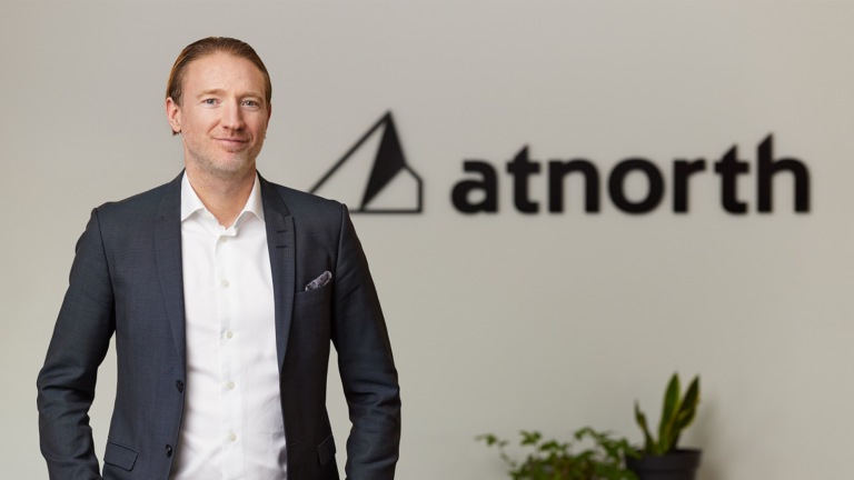 A man in a suit stands in front of a wall with the “atnorth” logo; there are potted plants in the background.