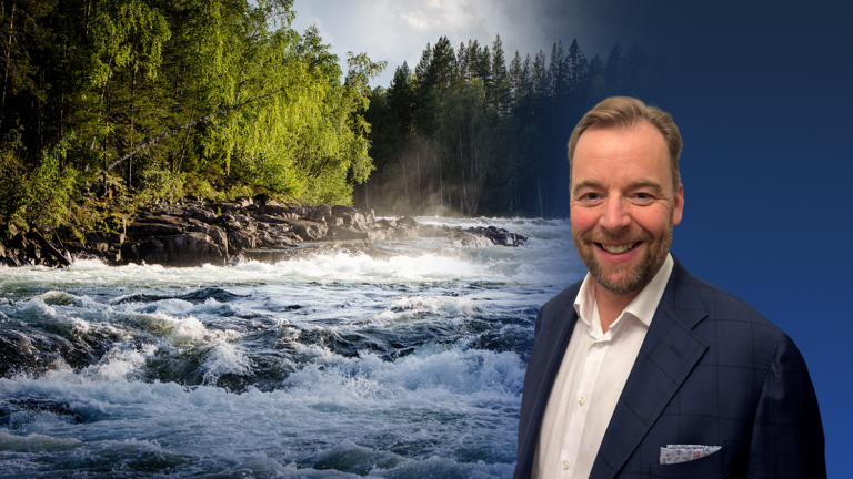 A man in a suit smiles in front of a background showing a fast-flowing river surrounded by trees under a partly cloudy sky, symbolizing the powerful flow and connectivity found in today’s AI-ready data centers.