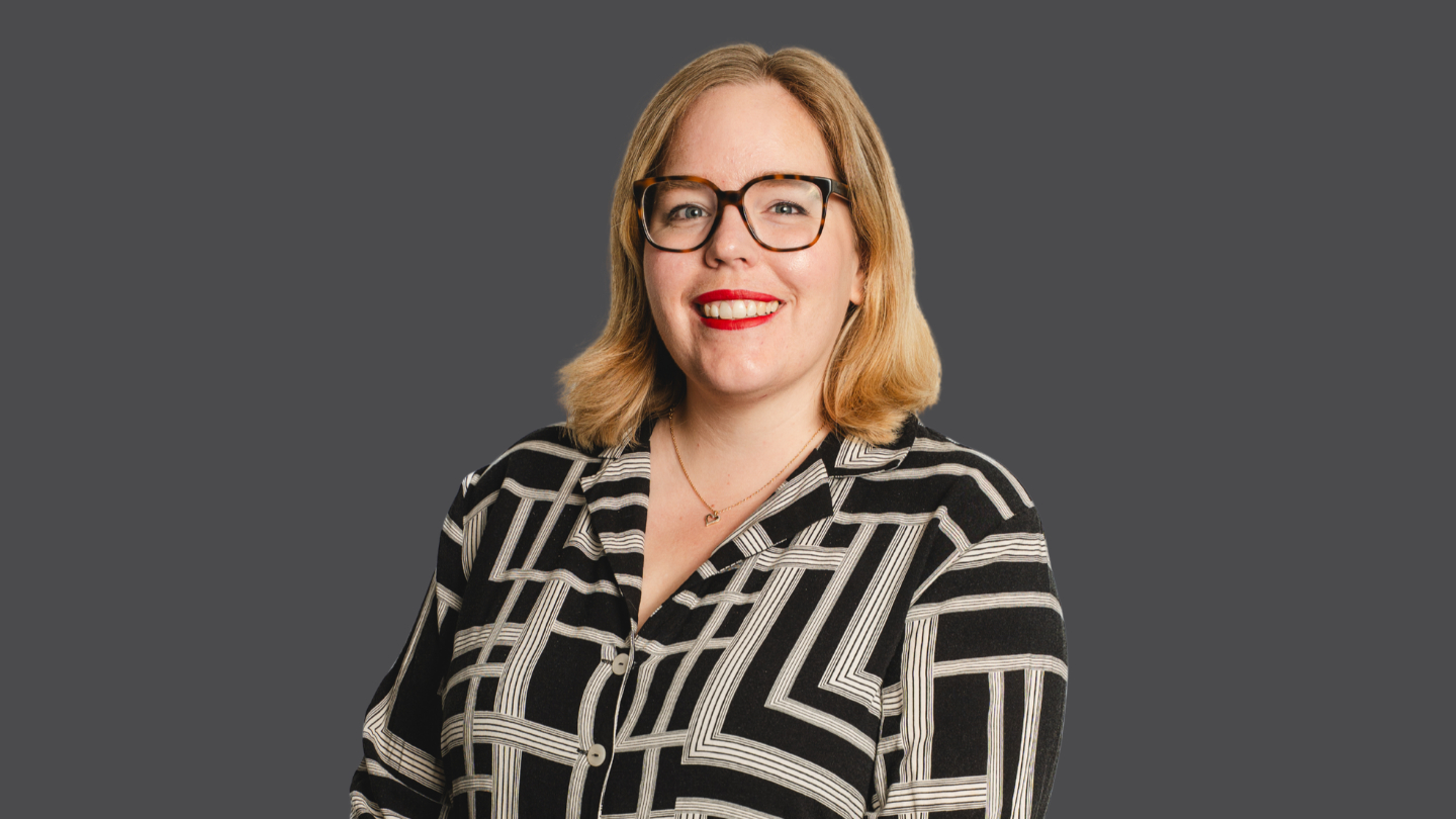 A woman with blonde hair and glasses wearing a black and white patterned shirt, smiling against a grey background.